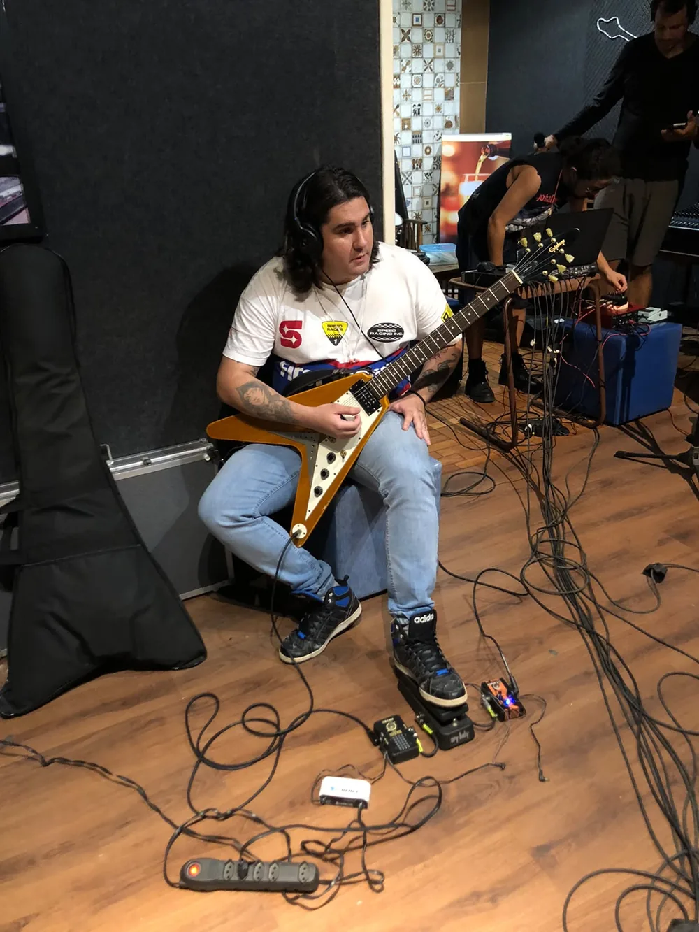 Estúdio: guitarrista sentado tocando uma guitarra Flying V, com cabos e pedais no chão